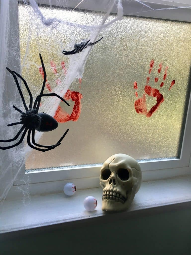spider webs and bloody handprints are covering a window with a skull on the window sill. 