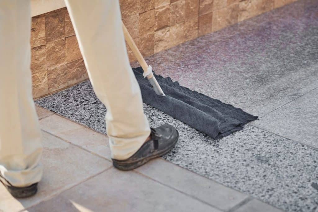 A person in khaki pants and black shoes cleaning a marble floor with a broom and a towel. 