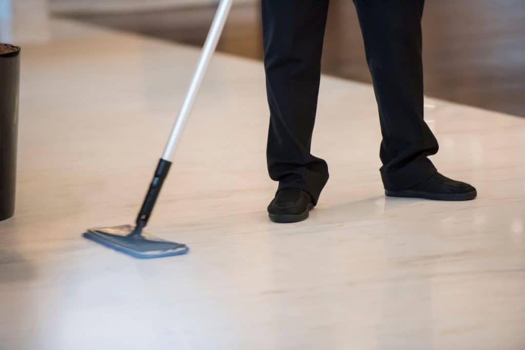 Person wearing black pants and shoes cleaning a marble floor with a dust mop. 