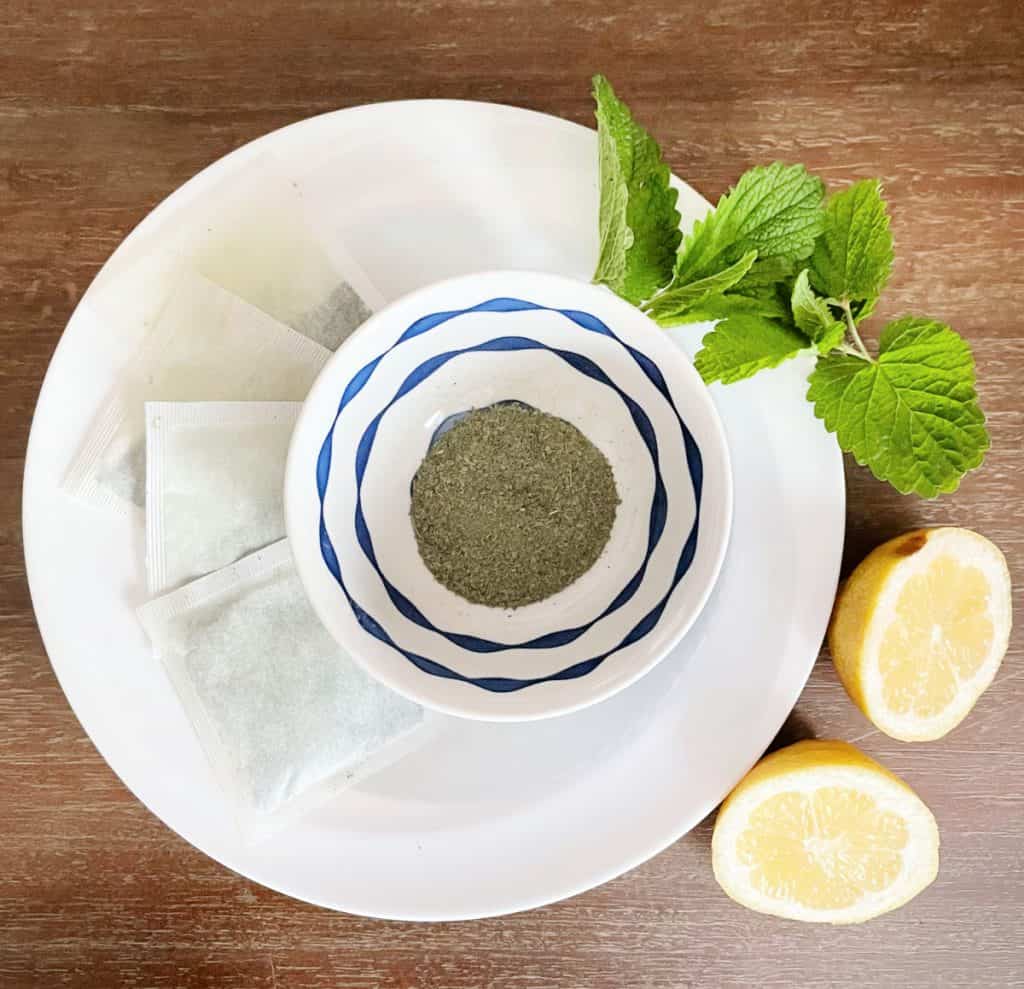 Bowl of loose green tea on a white plate with tea bags, lemon wedges and herb garnishes. 