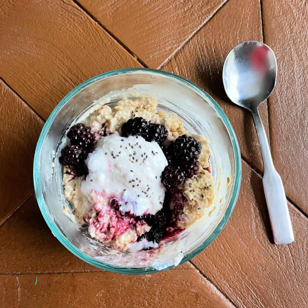 Overnight oats with protein powder in a glass bowl topped with blackberries, greek yogurt and chia seeds. 