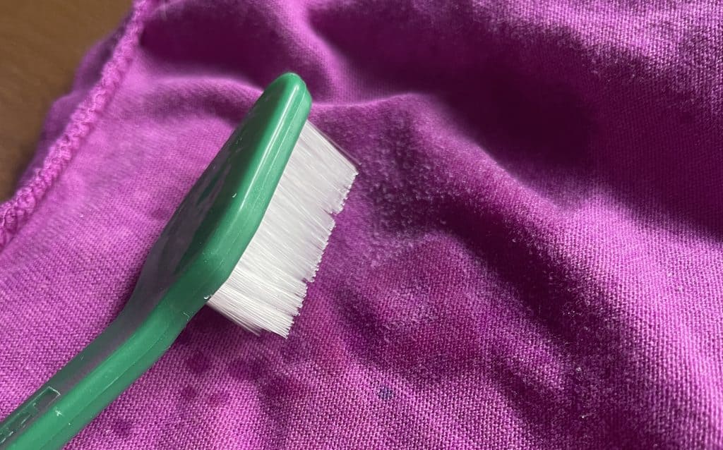 An old toothbrush gently scrubbing a t-shirt to get rid of chapstick from clothing. 