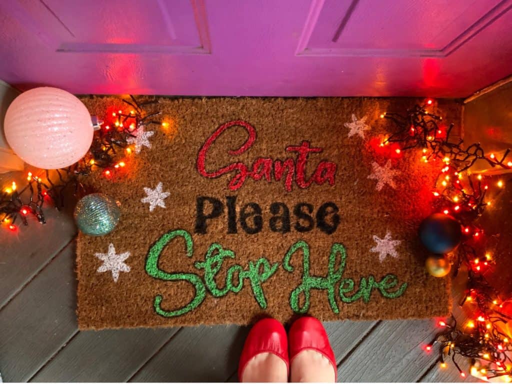 DIY doormat on a front porch with Christmas lights and ornaments. 