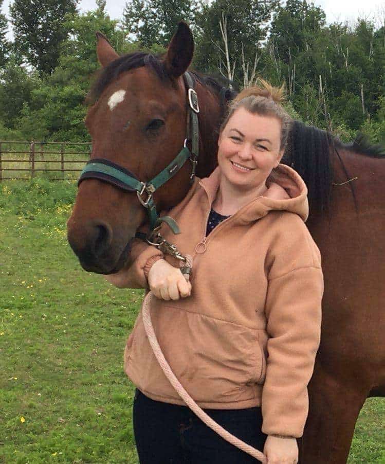 Woman holding the reigns of a brown horse in a grass pasture.